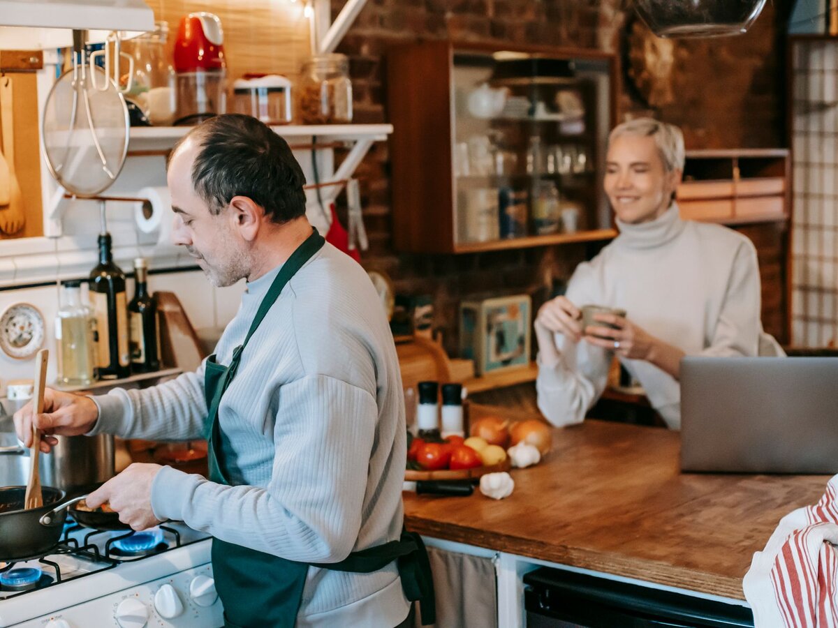 A man smiling and stirring a pot on the stove while a woman watches from an island counter