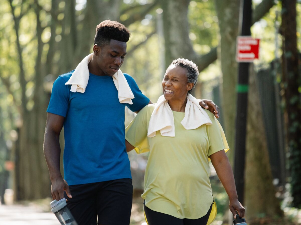 An older woman and a younger man walking and talking outdoors with towels around their necks.