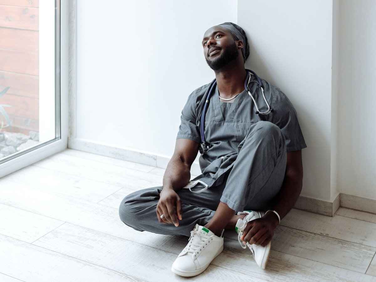A male nurse in scrubs sitting on the floor leaning against a wall and looking upward thoughtfully