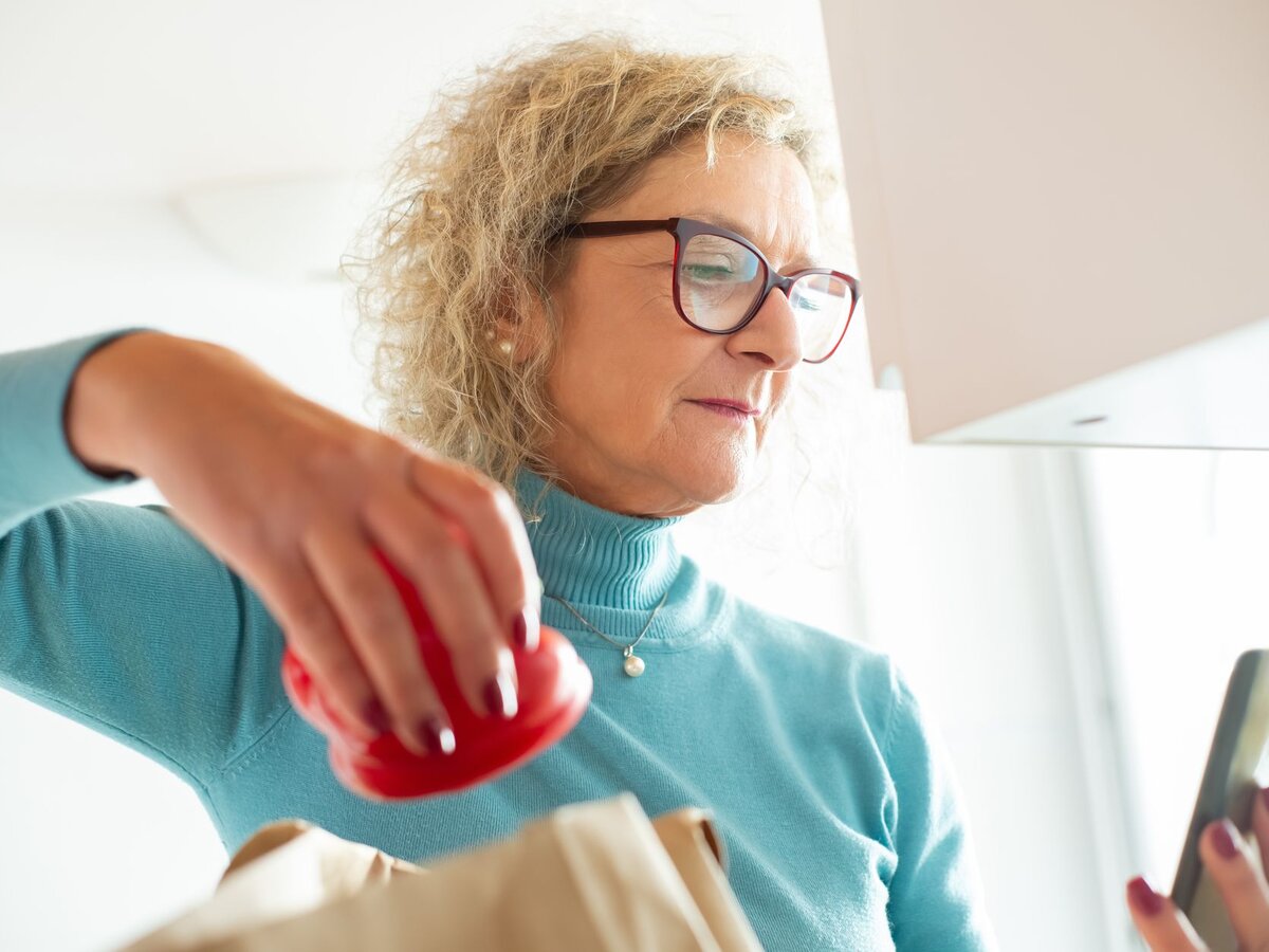 An older woman in glasses unpacking groceries in a bright kitchen.