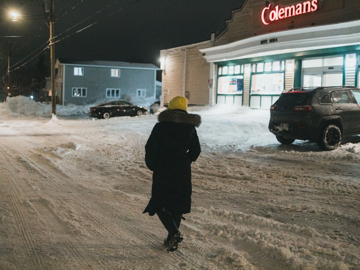 A woman walking alone down a snowy street at dusk near a storefront.