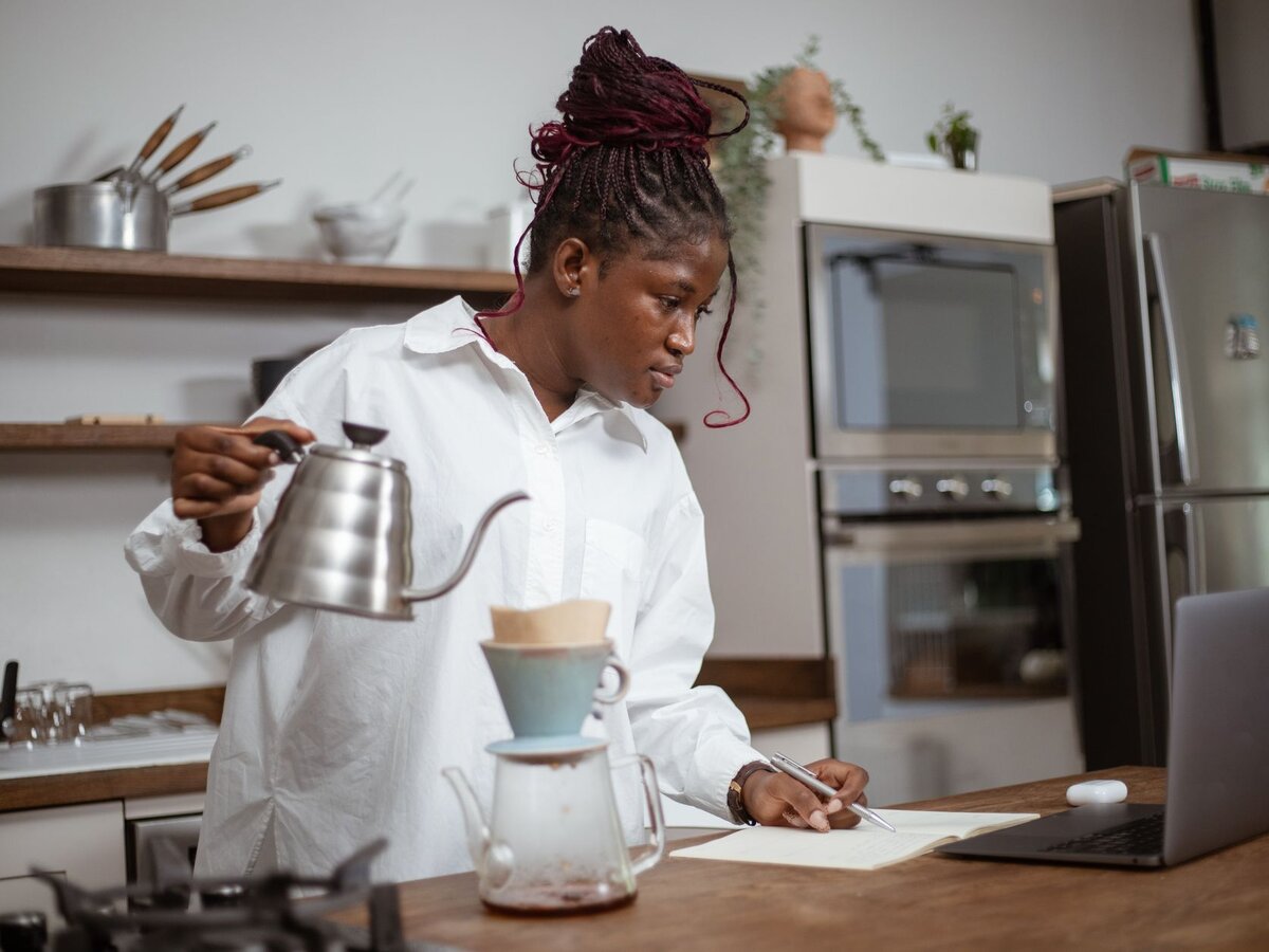 A woman pouring coffee into a pour-over filter while writing notes in a notebook next to a laptop.