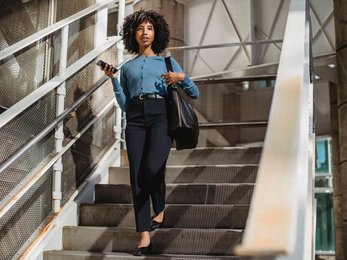 A woman carrying a bag walks down outdoor stairs in the sunlight.