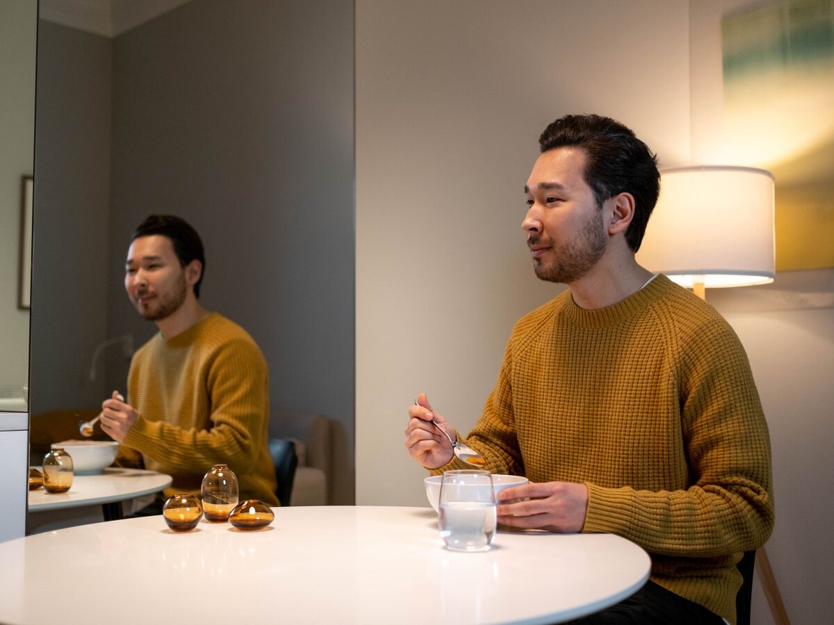 A man in a yellow sweater eating a simple meal at a white table indoors.